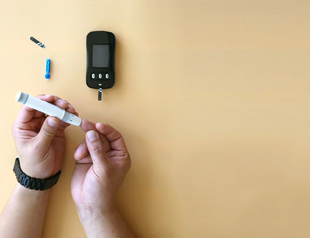 A person sitting at a kitchen table checking a continuous glucose monitor on their arm while reviewing notes in a diabetes logbook, morning coffee nearby